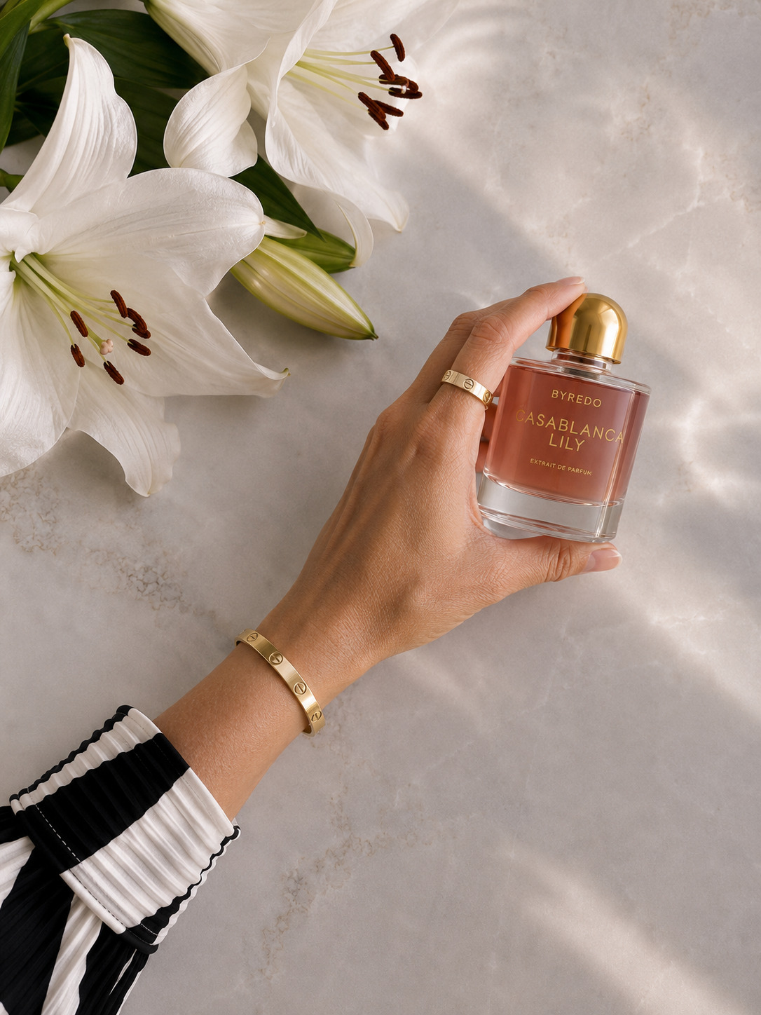Hand holding Byredo Casablanca Lily perfume beside white lilies on pale stone