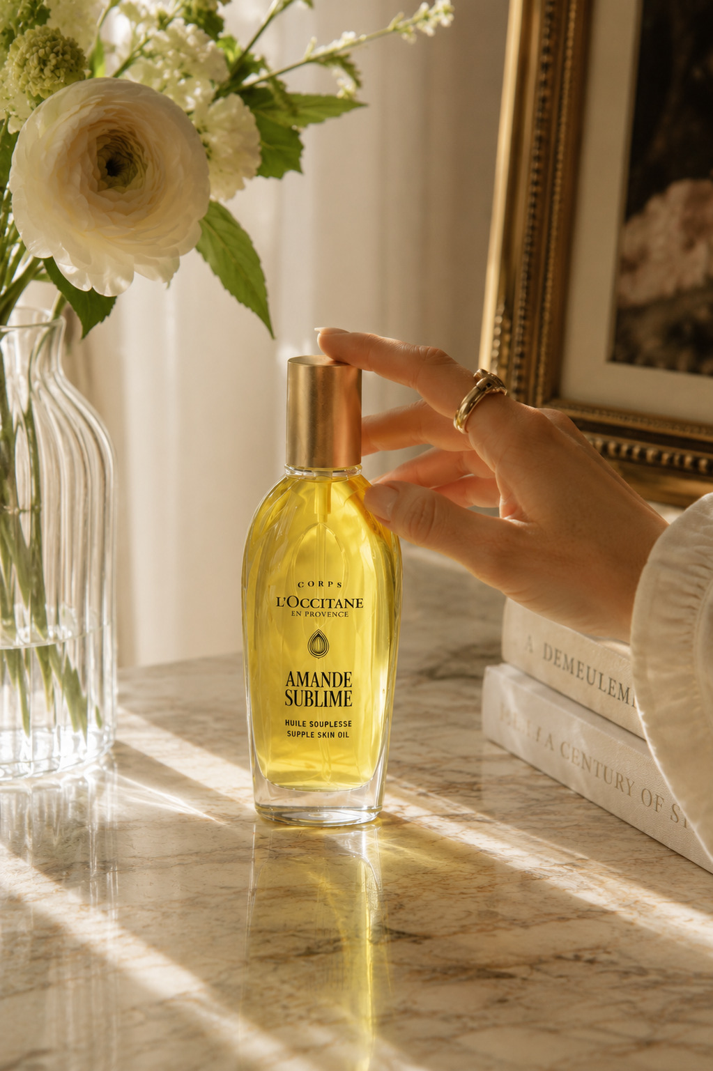 Hand reaching toward L'Occitane almond oil beside white flowers in a sunlit interior