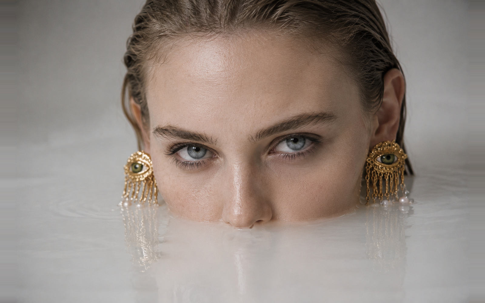 Editorial beauty portrait of a woman partially submerged in milky water wearing sculptural gold earrings