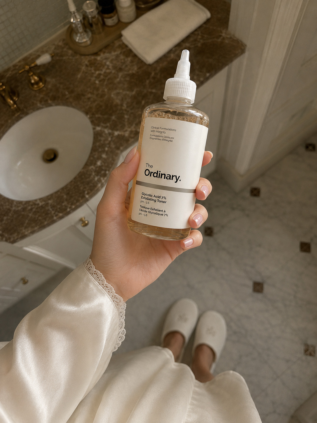 Hand holding The Ordinary skincare toner in a warm marble bathroom